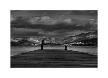 black and white photo of the end of a wooden jetty overlooking a lake with mountains and dark clouds in the background
